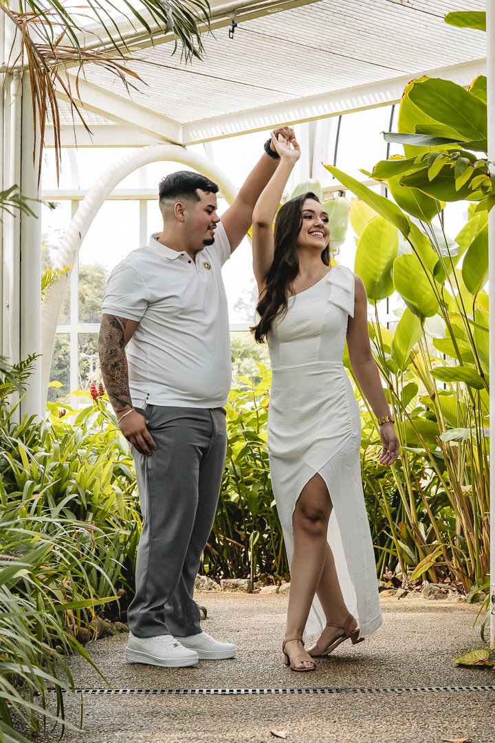 O noivo Gabriel segura no alto a mão da noiva Júlia enquanto ela roda numa bela dança no meio das plantas da estufa do Jardim Botânico de Curitiba