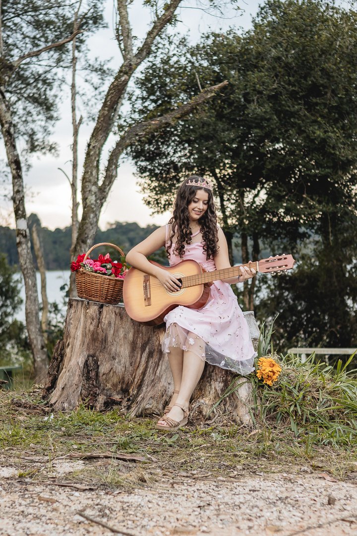 Luana, a debutante, está em um bosque usando um vestido até os joelhos cor-de-rosa com um voil bordado, sentada em um tronco de uma árvore cortada tocando violão. Ao seu lado direito uma cesta com diversas flores vermelhas e cor-de-rosa, e do outro lado u