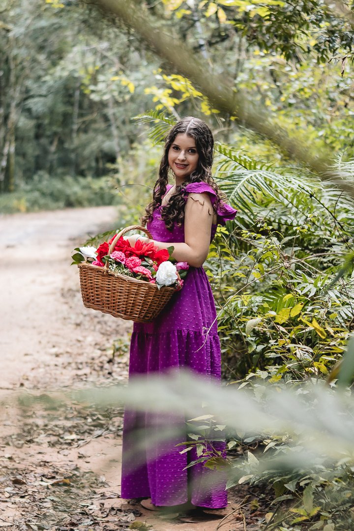 A debutante Luana em uma estrada de terra com bastante folhas nas bordas, usando um vestido roxo e segurando uma cesta cheia de flores coloridas
