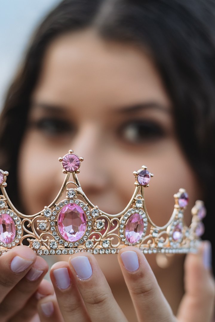 Luana, a debutante desfocada ao fundo e em primeiro plano ela segura uma bela coroa dourada com pedras cor-de-rosa