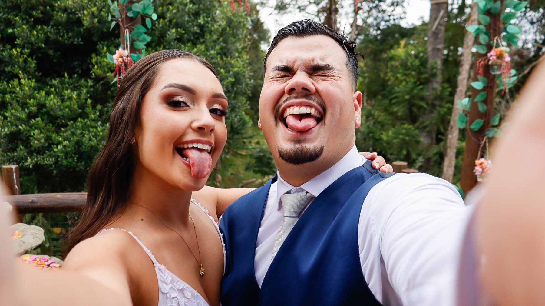 Os noivos sorrindo e mostrando a língua enquanto seguram a câmera para uma selfie.