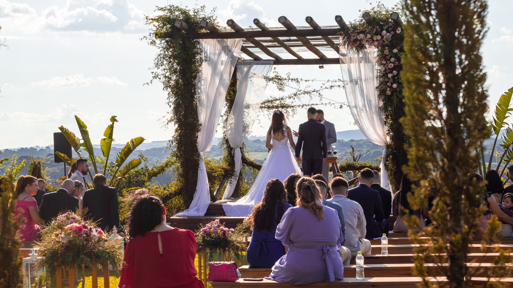 A cerimônia vista de costas. Os convidados sentados e ao fundo os noivos em pé no altar.